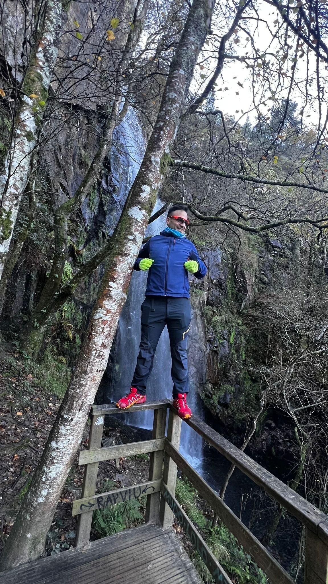 A person standing on a wooden bridge in a forested area with a waterfall in the background.