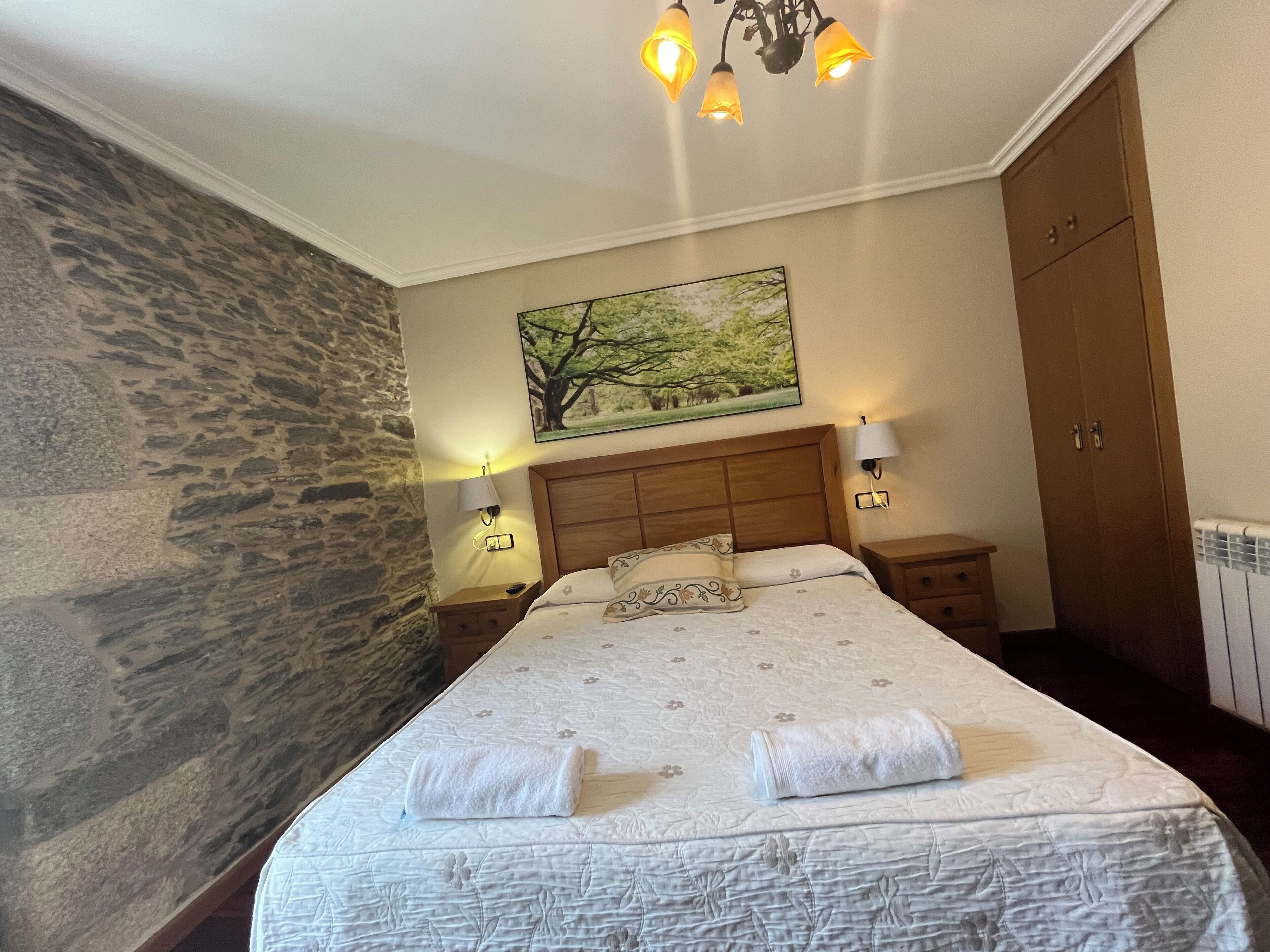 A bedroom with a stone wall, a wooden headboard, a painting of trees, a bed with a floral bedspread, and two bedside lamps.