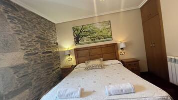A bedroom with a stone wall, a wooden headboard, a painting of trees, a bed with a floral bedspread, and two bedside lamps.