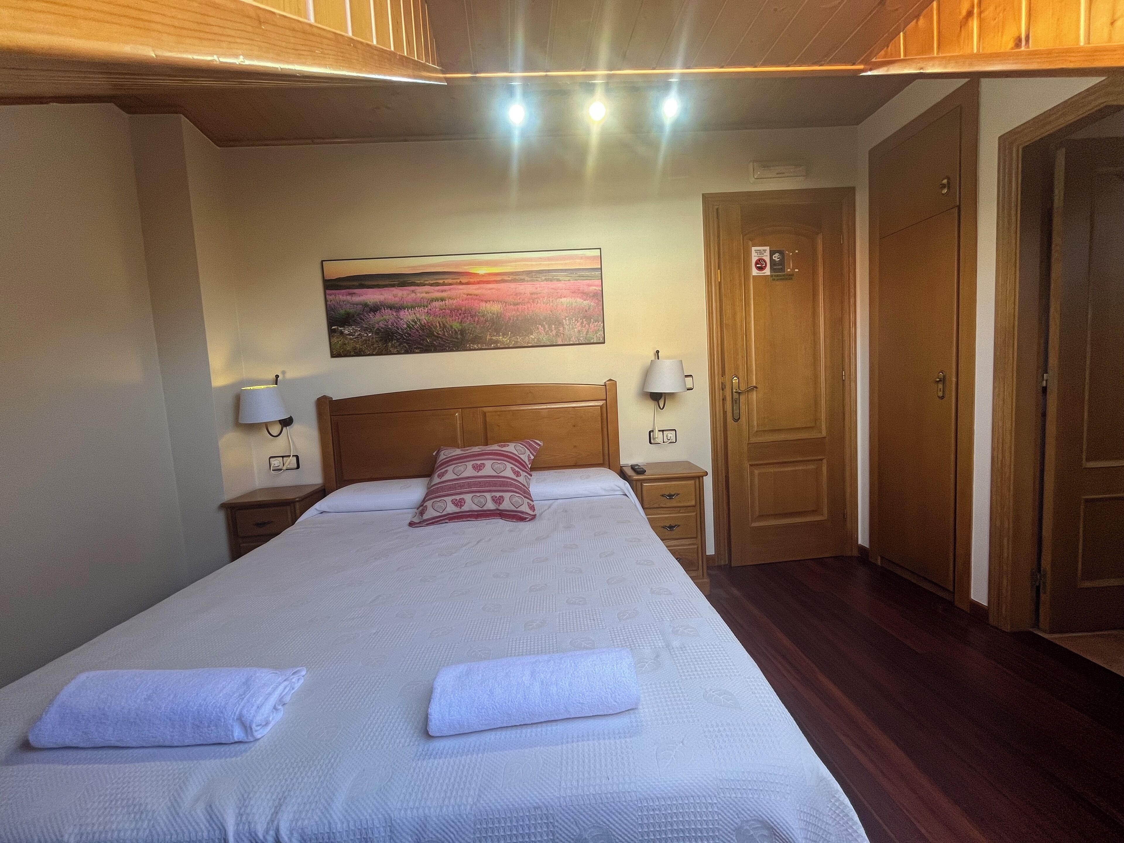 Een netjes opgemaakt bed met een kussen met patroon, een bedlampje en een schilderij van een zonsondergang aan de muur.