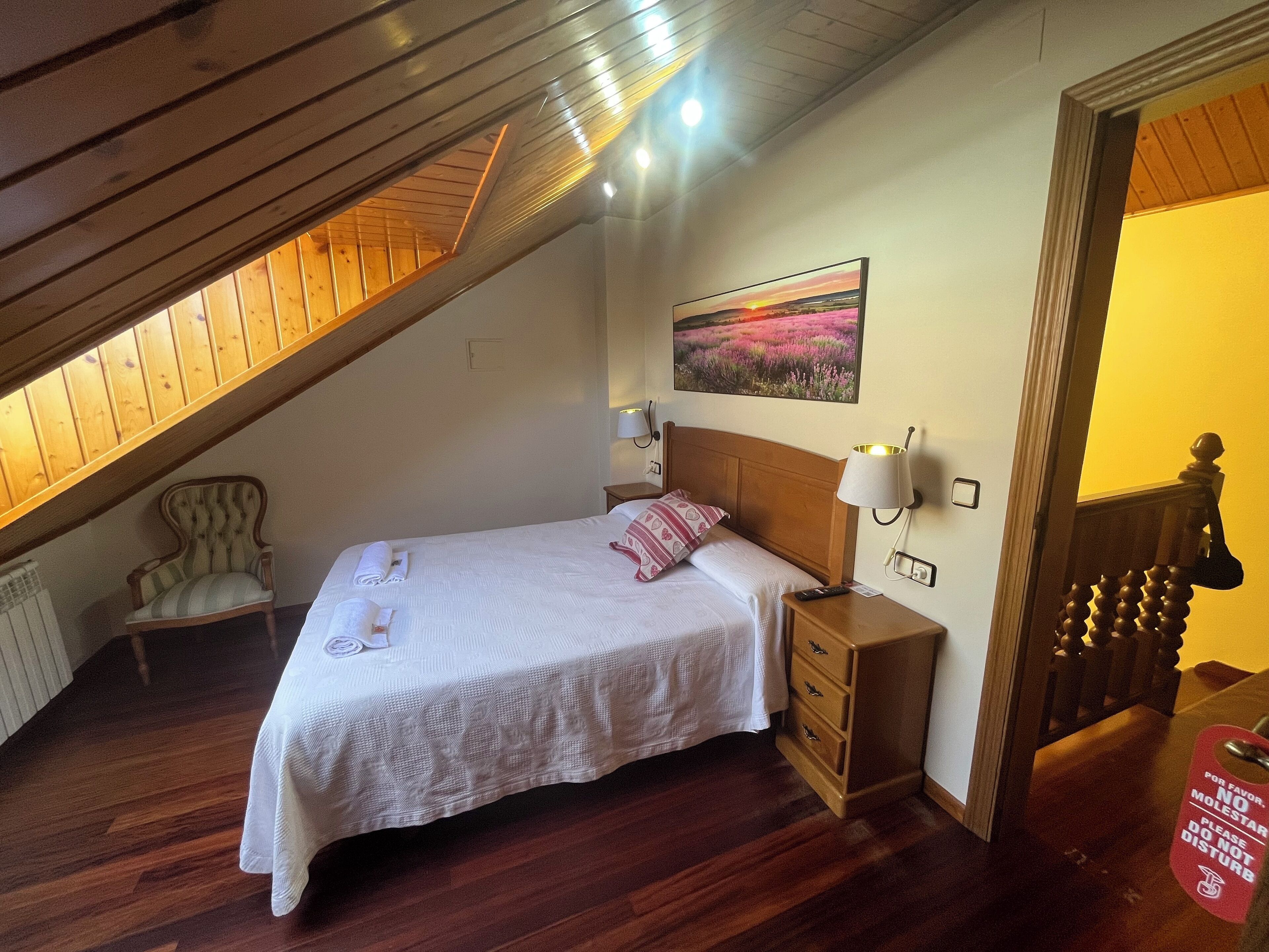 Un dormitorio con techo de madera, una cama con ropa de cama blanca, una mesita de noche, una silla y un cuadro en la pared.