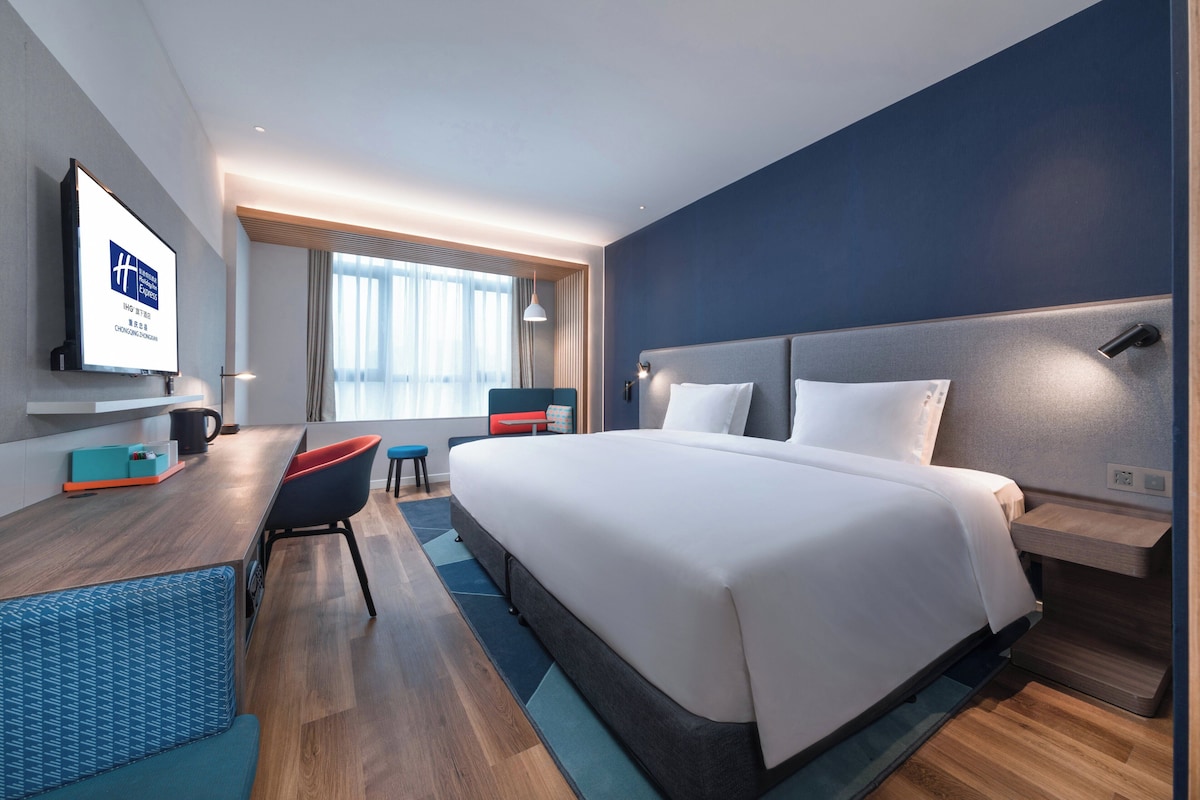 A modern hotel room with a large bed, a desk, a chair, and a television displaying a sign.
