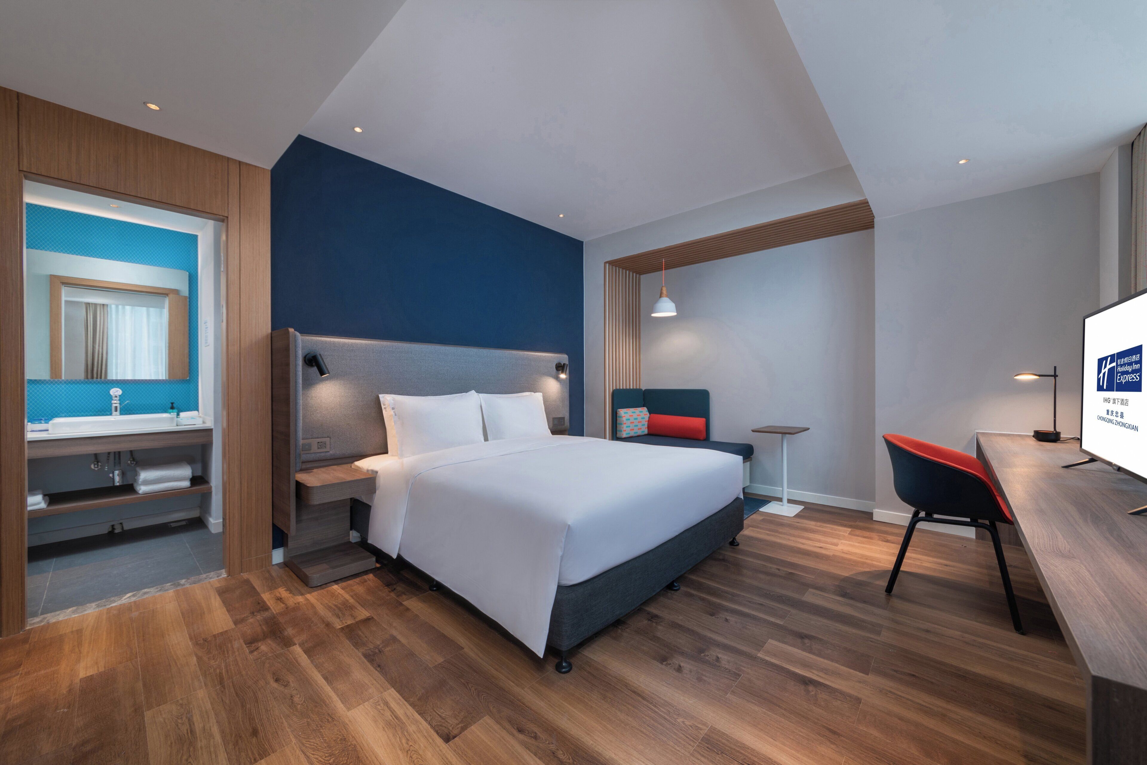 A modern hotel room with a large bed, a desk, a chair, and a bathroom with a mirror and sink.