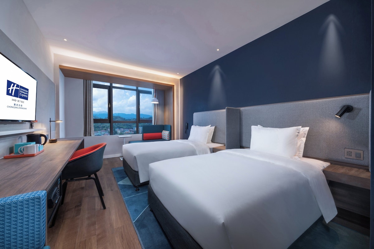 A hotel room with two beds, a desk, and a view of the city.