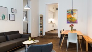 A modern living room with a sofa, a round table, and a dining area with chairs. There is a bookshelf with books and a painting on the wall. - BENSIMON apartments Charlottenburg (Berlin)