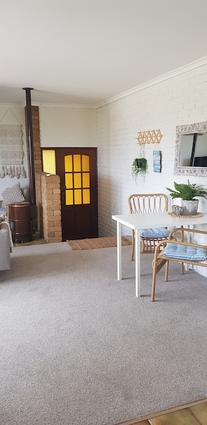 A cozy room with a white brick wall, a wooden door with a window, a white table with wicker chairs, and a small potted plant.