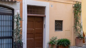 Une cour pavée de pierres, avec une porte à deux battants de couleur brune, des plantes en pot et une petite fenêtre.