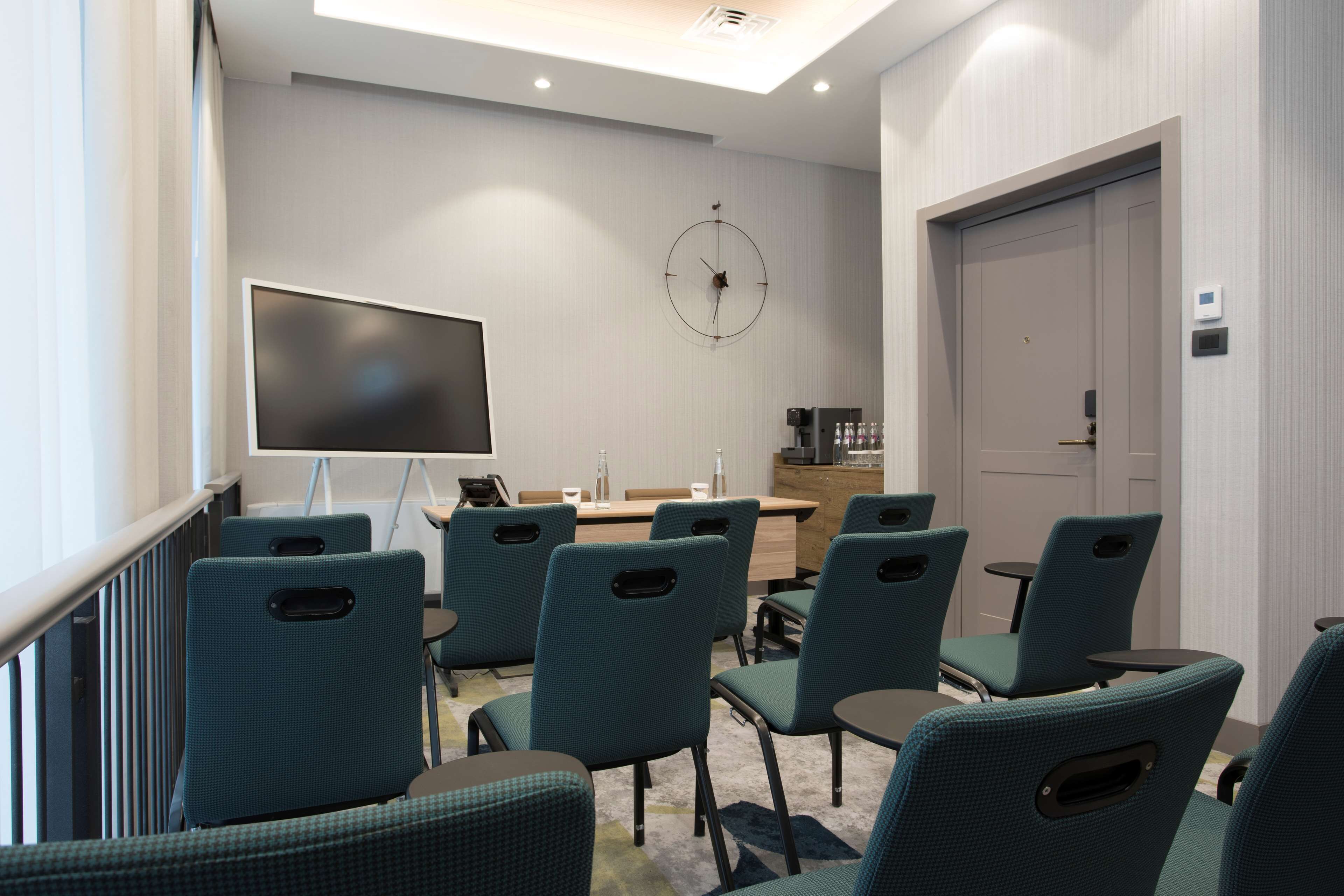 A conference room with teal chairs, a large screen, a clock, and a coffee machine.