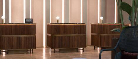 A hotel reception area with three wooden reception desks, each with a lit sign and a spherical light fixture.