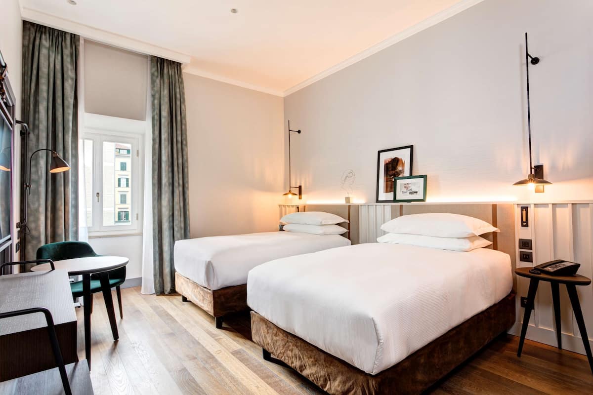 A hotel room with two single beds, a small table with a chair, a desk, and a window with curtains.