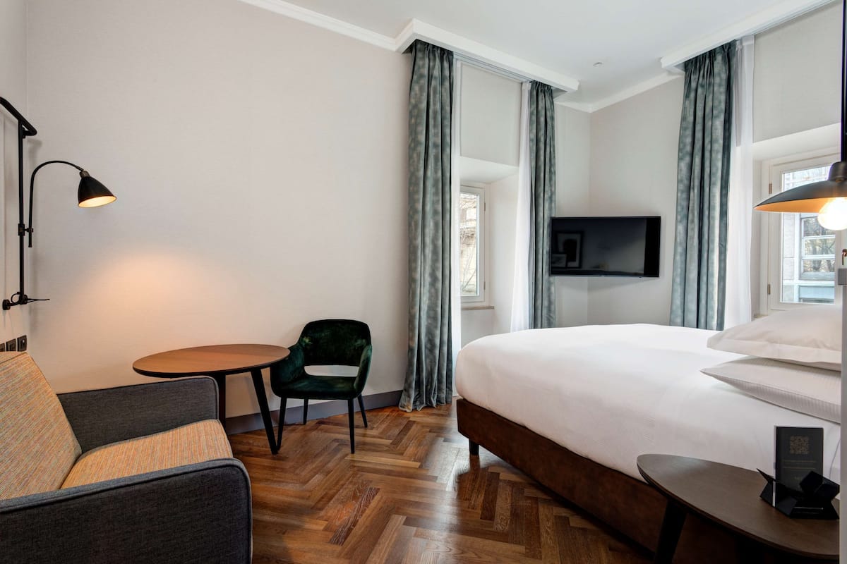 A hotel room with a bed, a small table, a chair, a desk, a TV, and curtains.