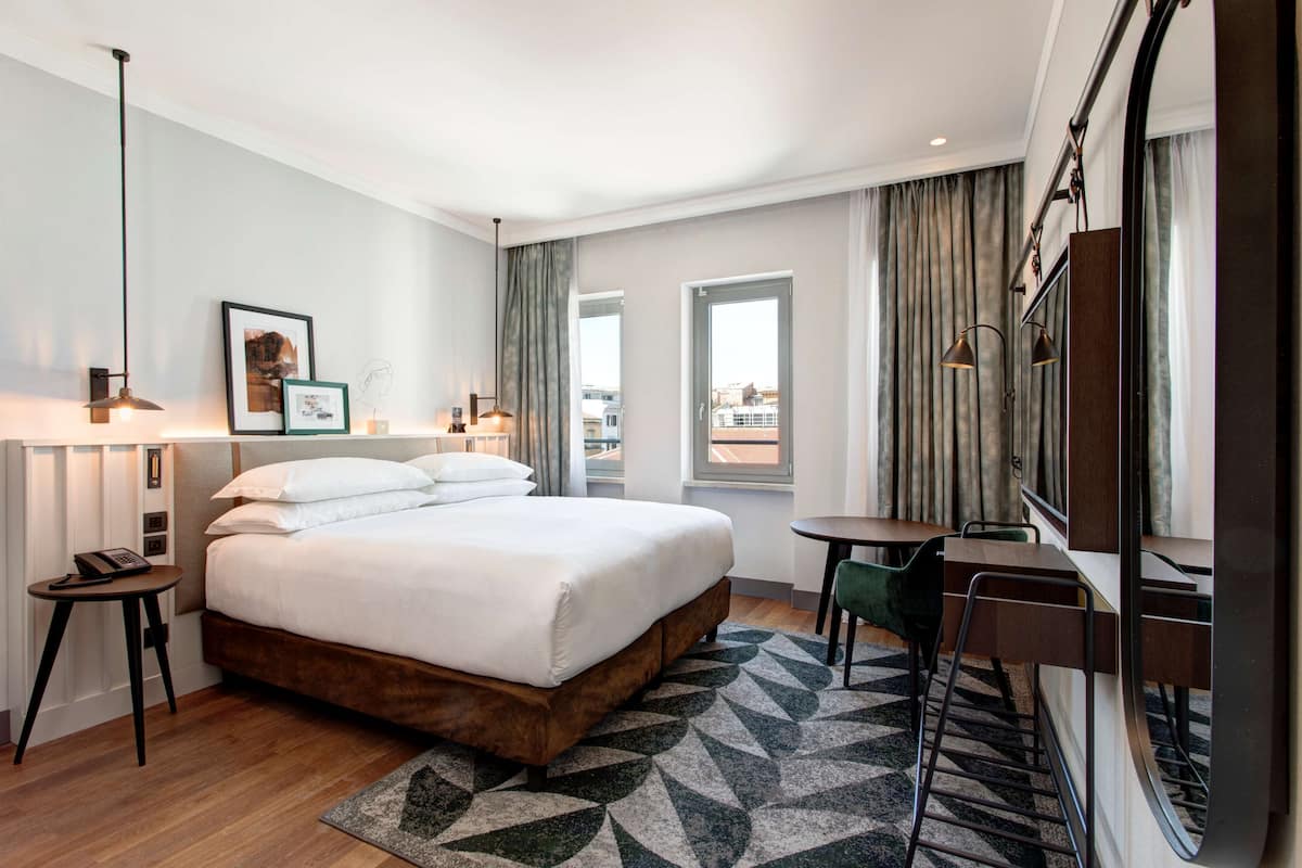 A hotel room with a large bed, a desk, a chair, a lamp, a picture on the wall, and a window with curtains.