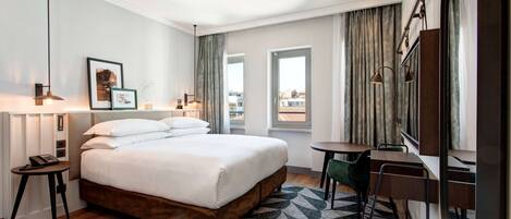 A hotel room with a large bed, a desk, a chair, a lamp, a picture on the wall, and a window with curtains.