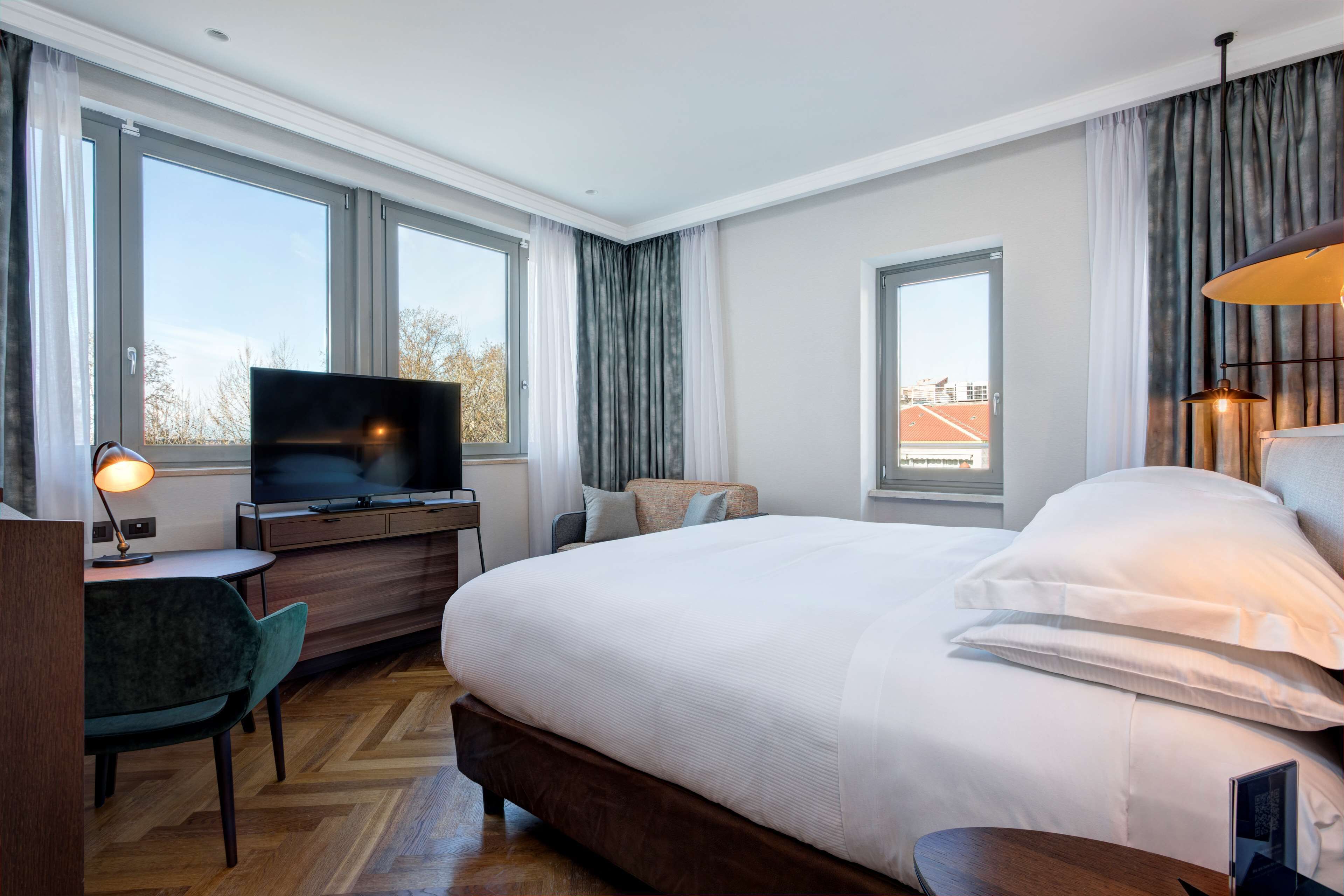 A hotel room with a large bed, a desk with a computer, a television, and a view of the city through large windows.