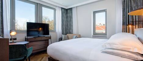 A hotel room with a large bed, a desk with a computer, a television, and a view of the city through large windows.