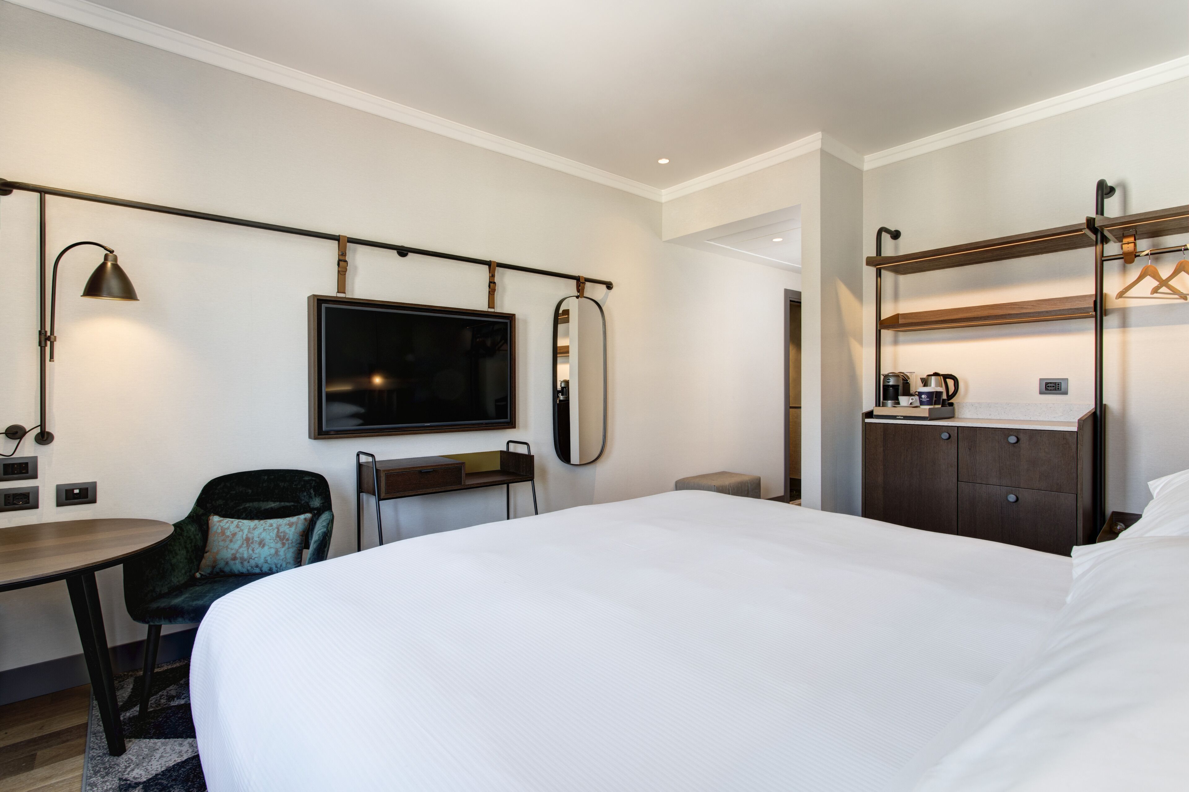 A hotel room with a large bed, a desk with a chair, a television, a mirror, a wardrobe, and a shelf with a kettle.