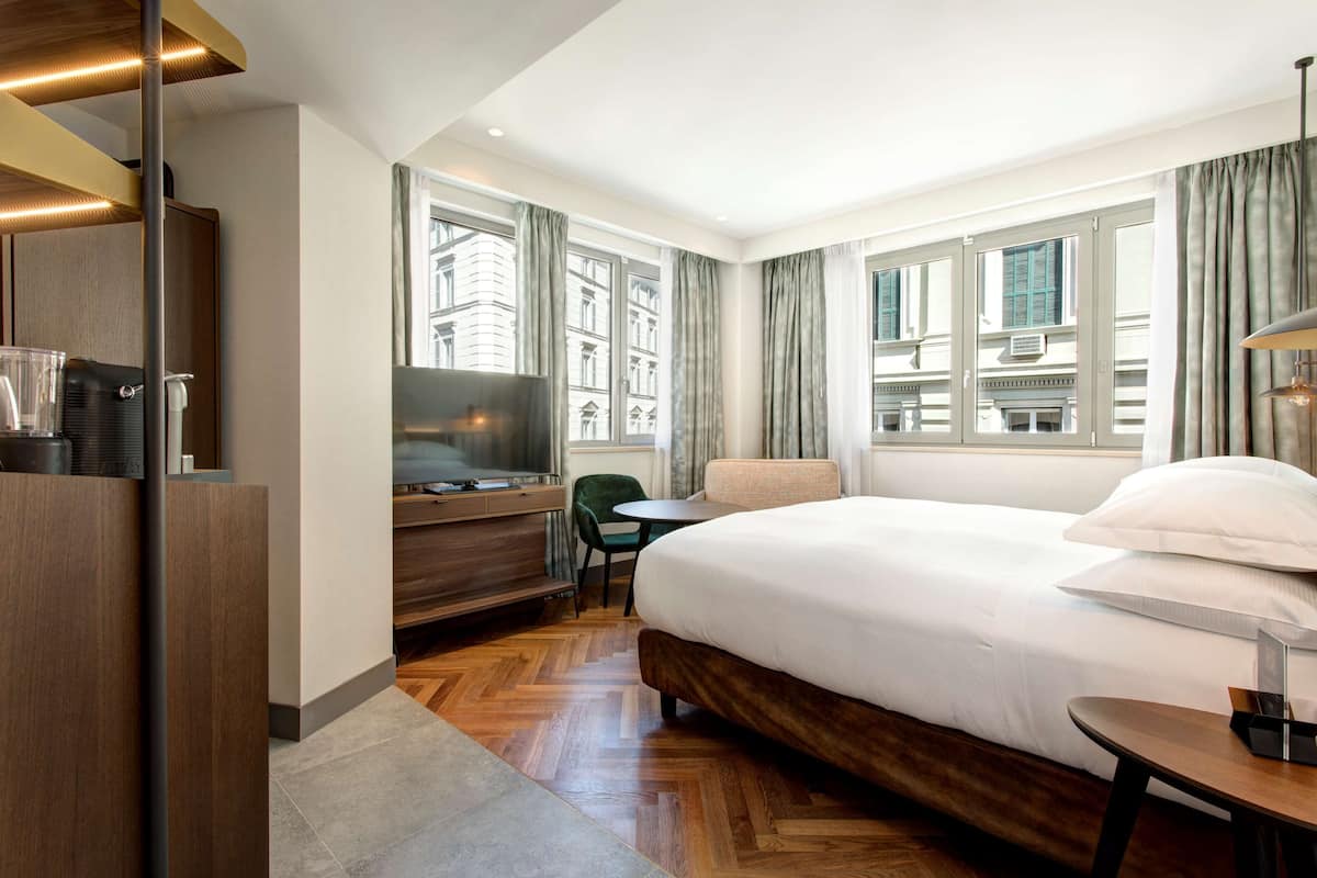A hotel room with a large bed, a desk with a TV, a chair, and a view of buildings through the windows.