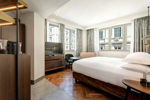 A hotel room with a large bed, a desk with a TV, a chair, and a view of buildings through the windows. - DoubleTree by Hilton Rome Monti (Rome)