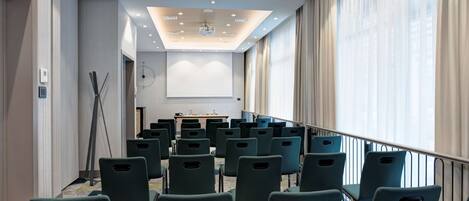 Un salón de conferencias con filas de sillas, una pantalla de proyector y una mesa para presentaciones.