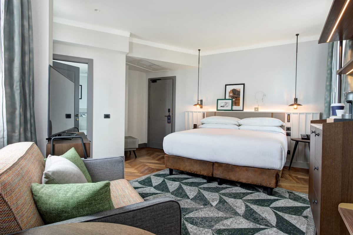 A modern hotel room with a large bed, a sofa, a desk, and a mirror.