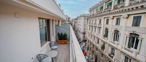 A narrow urban street with buildings on either side, a balcony with chairs and a plant, and a clear sky.