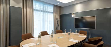 A conference room with a large table, brown chairs, a flat-screen TV, and a window with curtains.