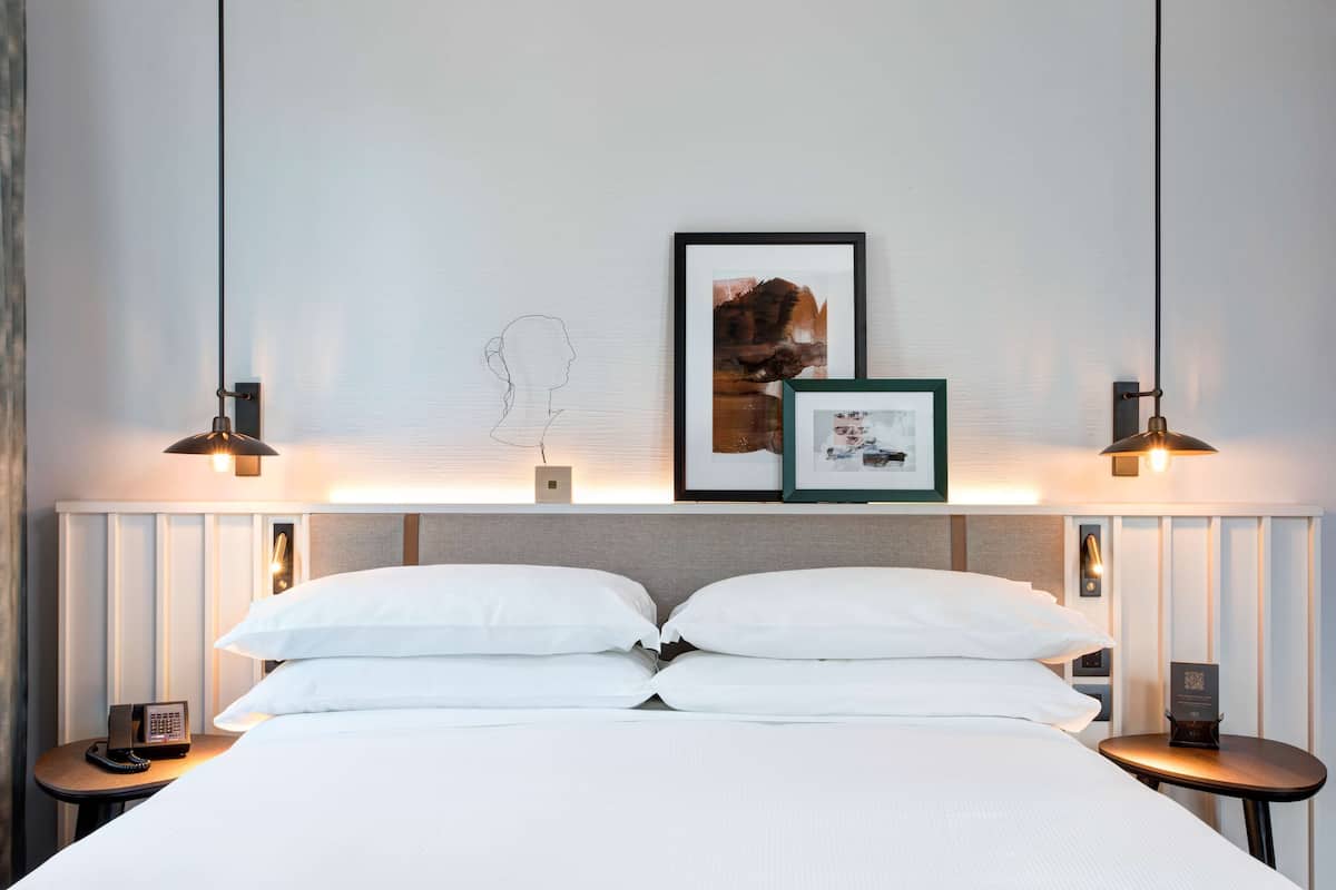 Una cama bien hecha con sábanas blancas, dos mesitas de noche con lámparas y cuadros en la pared.