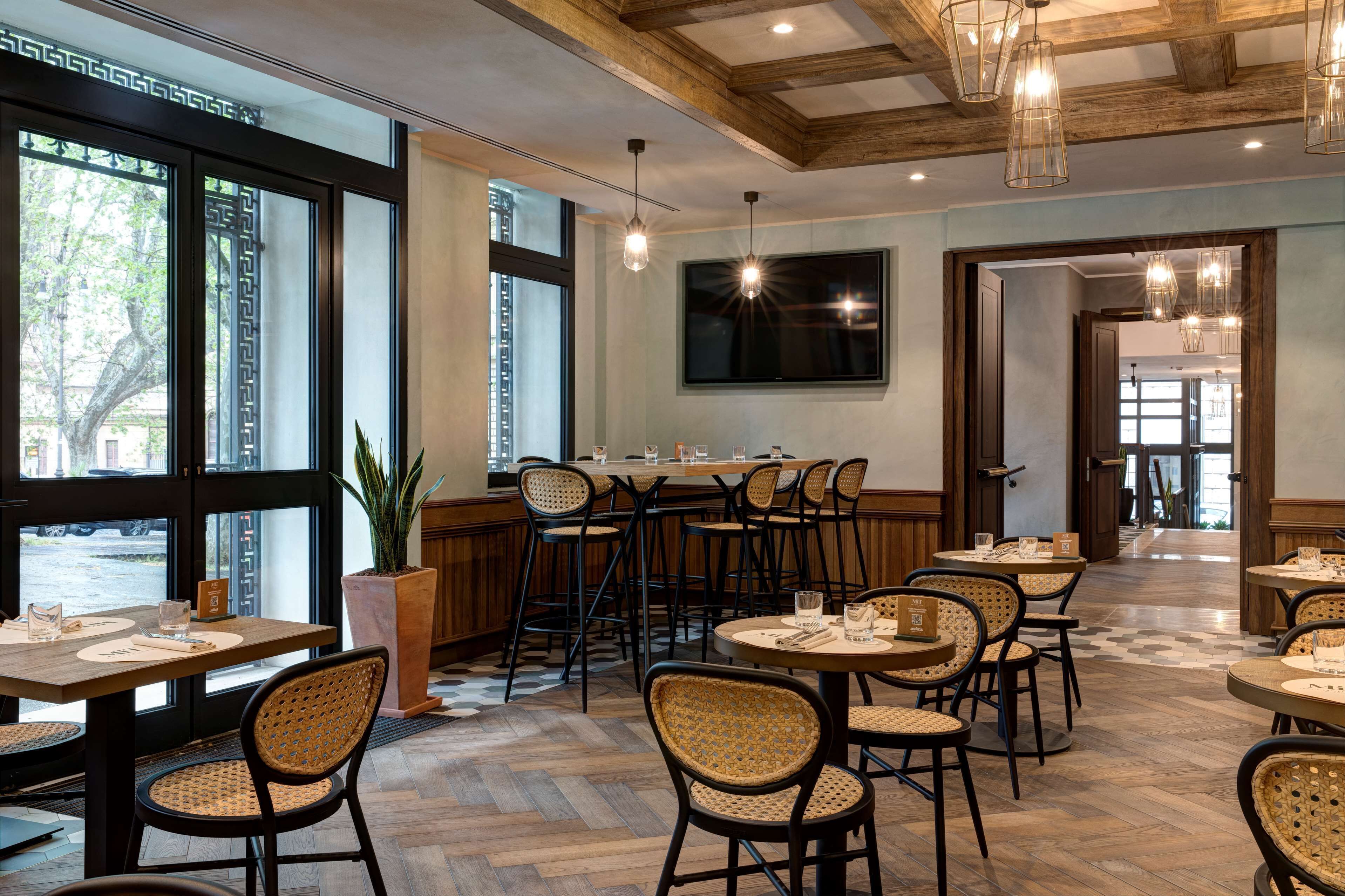 A restaurant with wooden beams, large windows, and wicker chairs. Tables are set with glasses and menus.