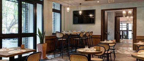 A restaurant with wooden beams, large windows, and wicker chairs. Tables are set with glasses and menus.