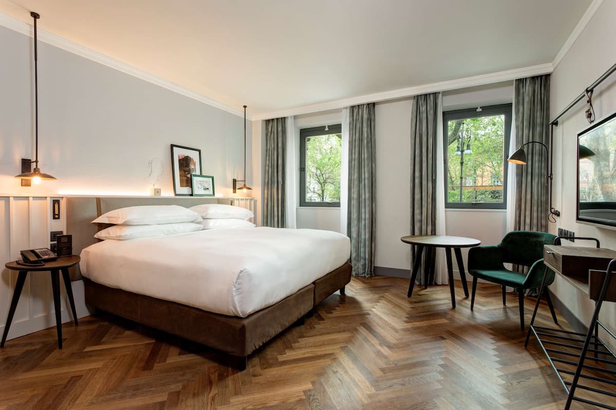 A hotel room with a large bed, a small round table, a green chair, and a desk with a chair. There are two windows with curtains, a lamp on the left, and a TV on the right.