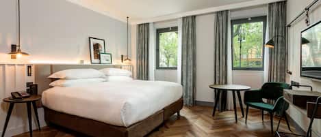 Une chambre d’hôtel avec un grand lit, une petite table ronde, une chaise verte et un bureau avec une chaise. Il y a deux fenêtres avec des rideaux, une lampe sur la gauche et un téléviseur sur la droite.