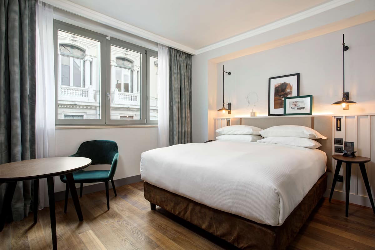 A hotel room with a large bed, a small table, a chair, and a window with curtains.