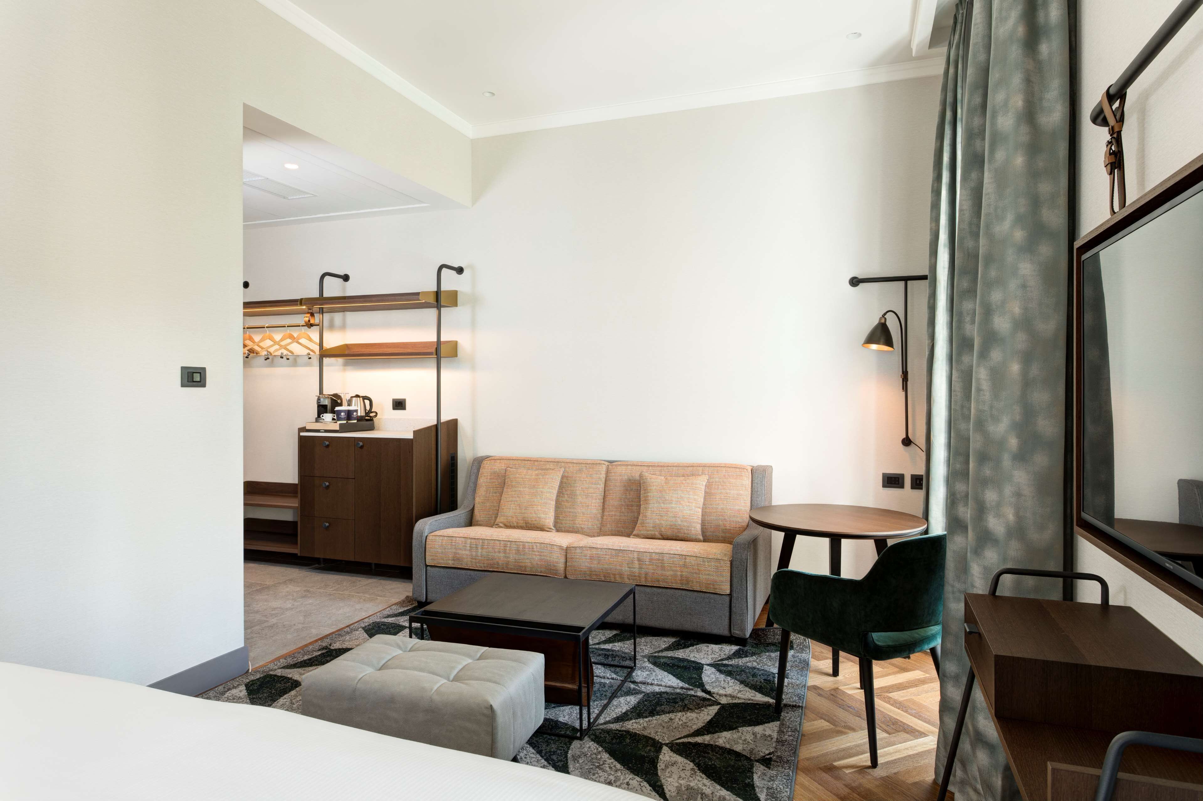 Une chambre d’hôtel moderne équipée d’un canapé, d’une petite table, d’une chaise, d’une table basse et d’un bureau. On aperçoit un coin cuisine au fond de la pièce.
