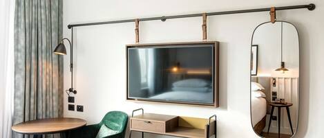 Habitación de hotel con cama, escritorio, silla, televisión, espejo y lámpara.