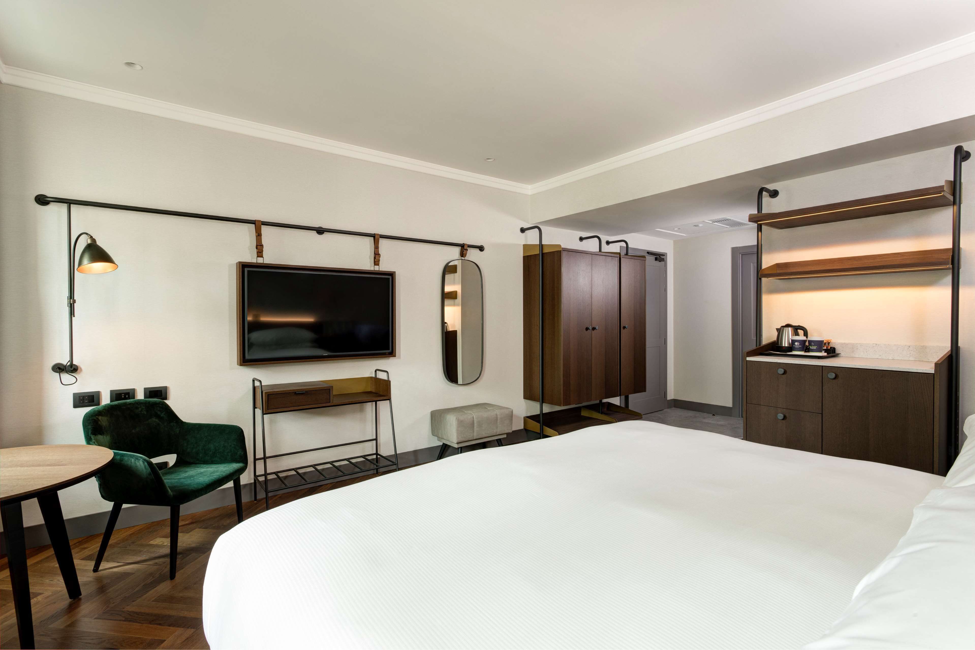 Una habitación de hotel moderna con una cama grande, un escritorio con silla, un televisor, un armario y una estantería con tetera.