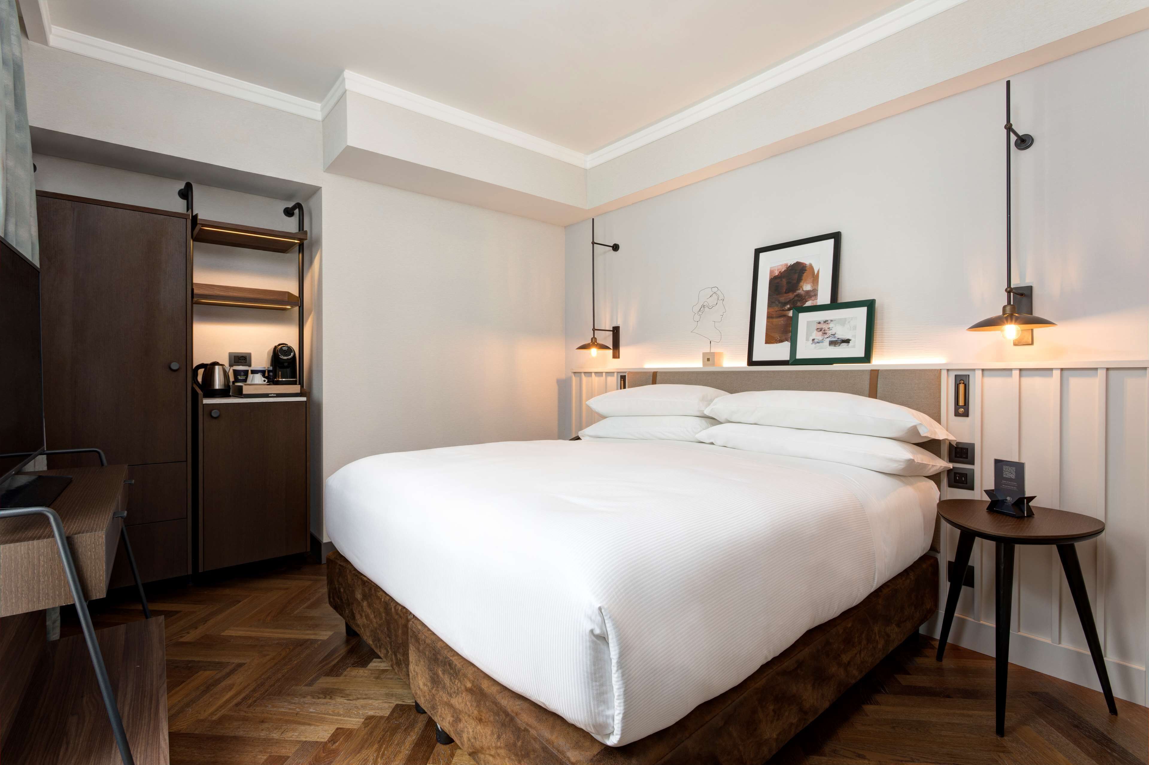 A neatly arranged hotel room with a large bed, a desk, a wardrobe, and a small table with a lamp.