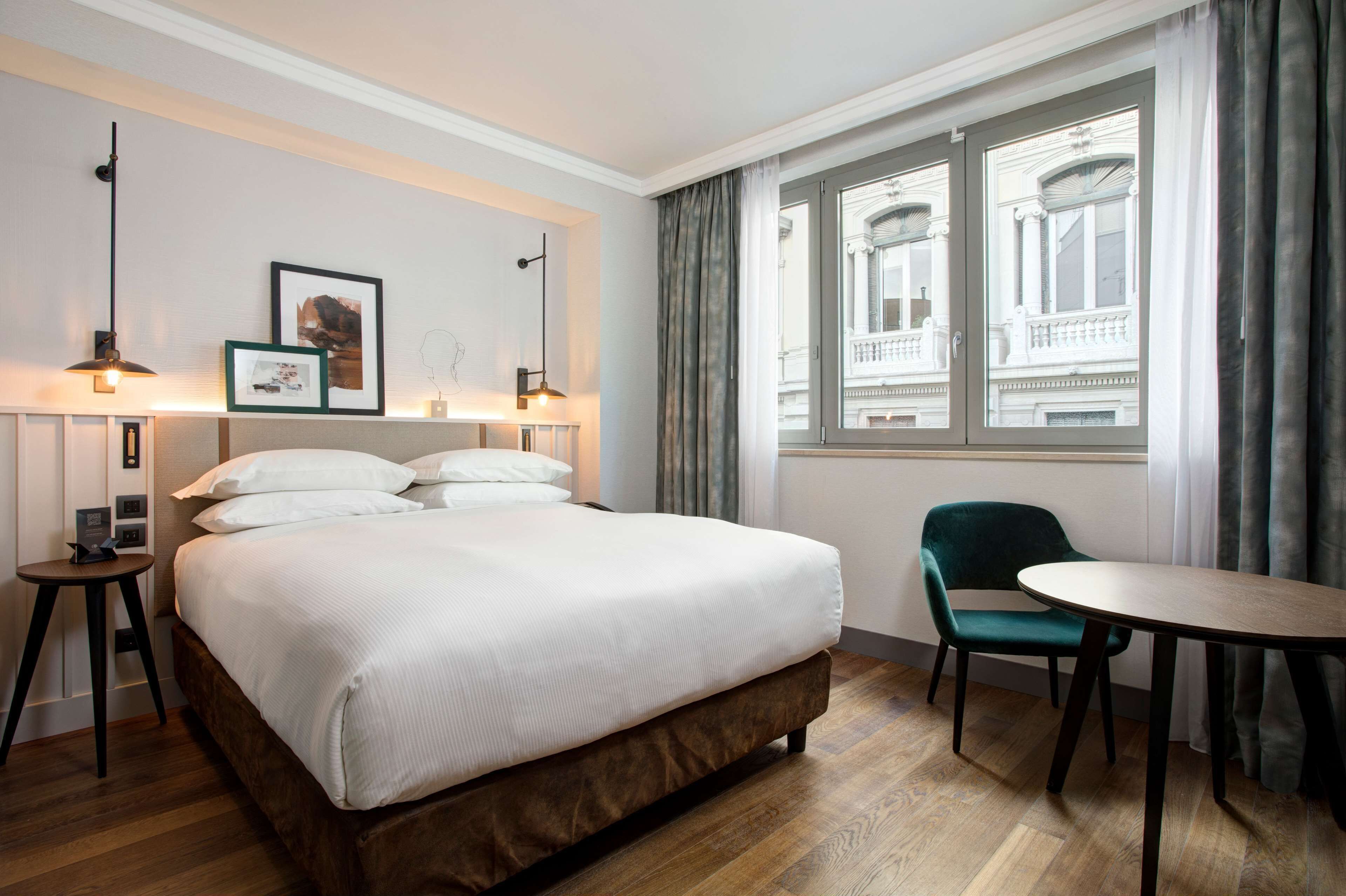 Une chambre d’hôtel avec un grand lit, une petite table, une chaise et une fenêtre donnant sur un immeuble.