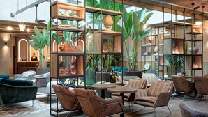 Un rebedor d'hotel modern amb un sostre alt, bigues al descobert i diverses opcions d'asseu-se, incloent sofàs i butaques. Hi ha grans finestres que deixen entrar llum natural, i l'espai està decorat amb plantes i diversos objectes decoratius sobre prestatgeries.