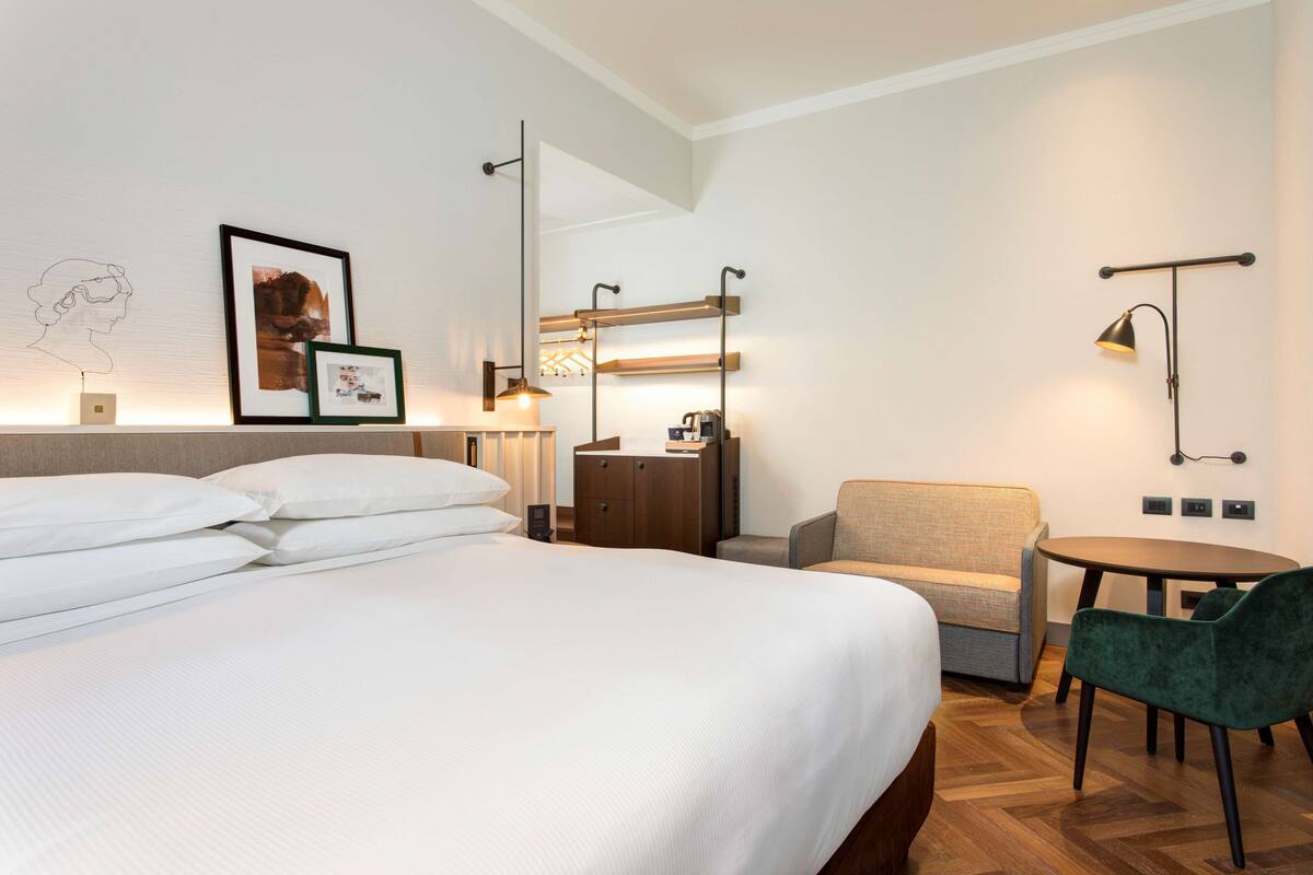 Habitación de hotel con una cama grande, un escritorio con silla, una mesita pequeña e iluminación en la pared.