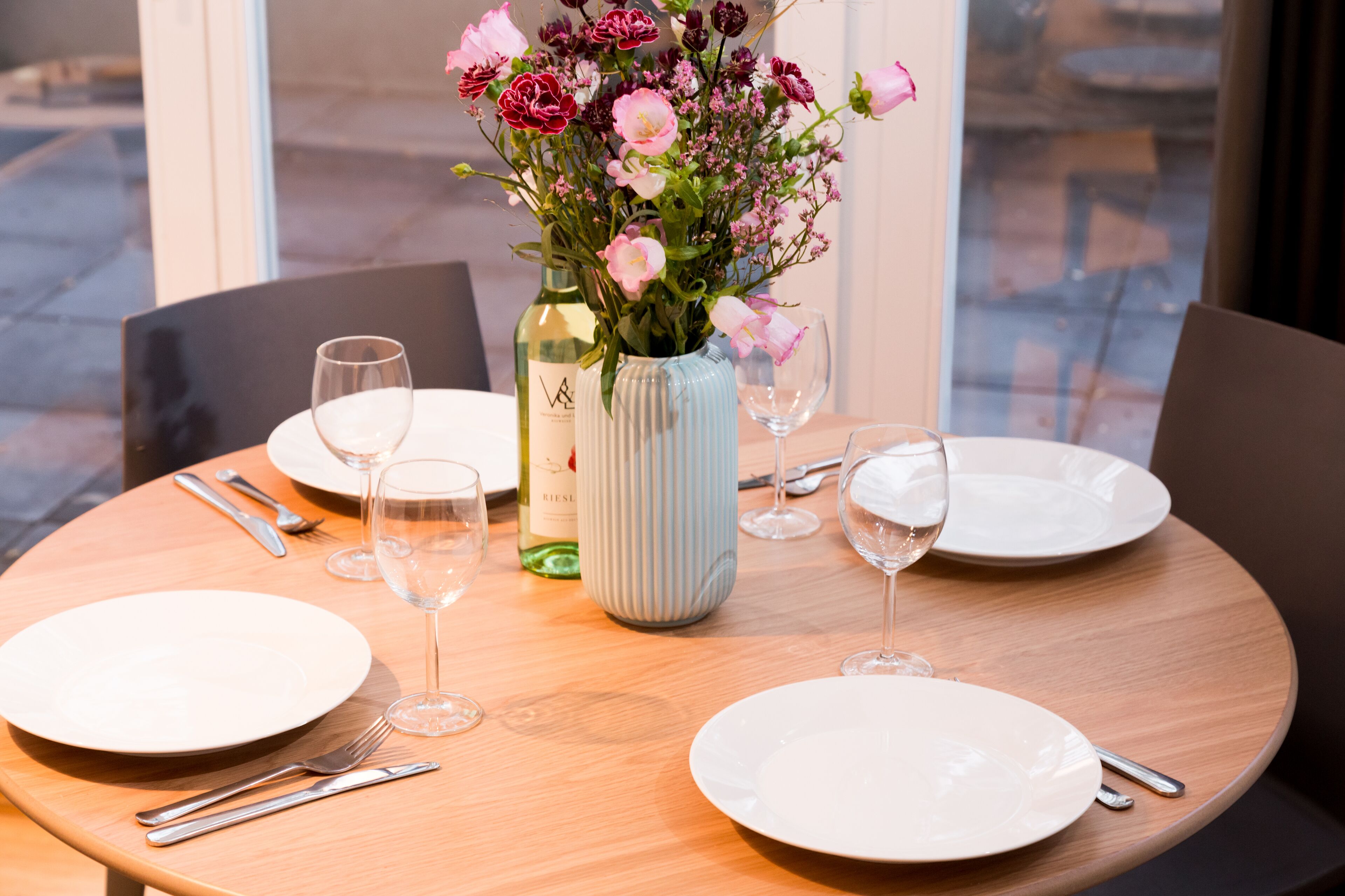 Una mesa de comedor dispuesta con platos, cubiertos, copas de vino y un florero con flores.