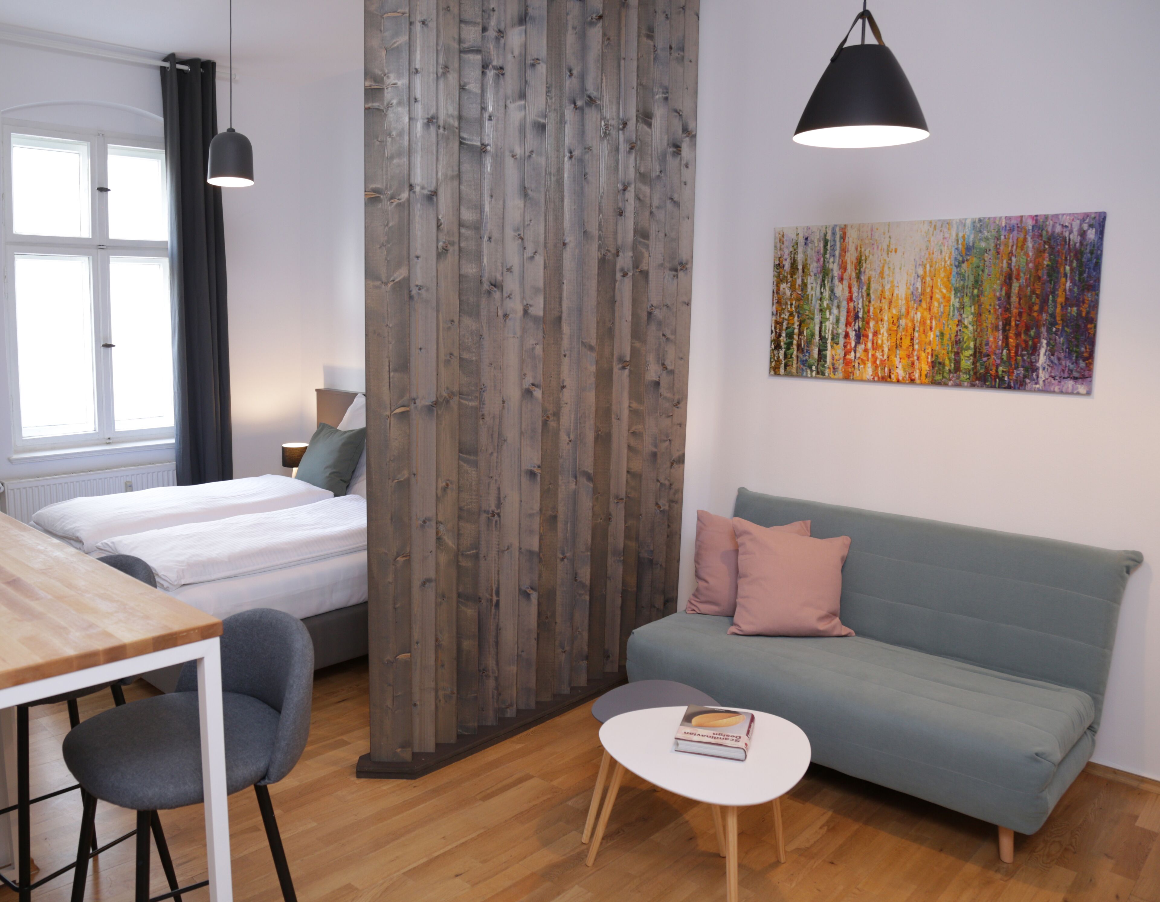 Quarto moderno com divisória de madeira, cama, sofá, mesa de jantar com cadeiras e uma pequena mesa redonda com um livro.