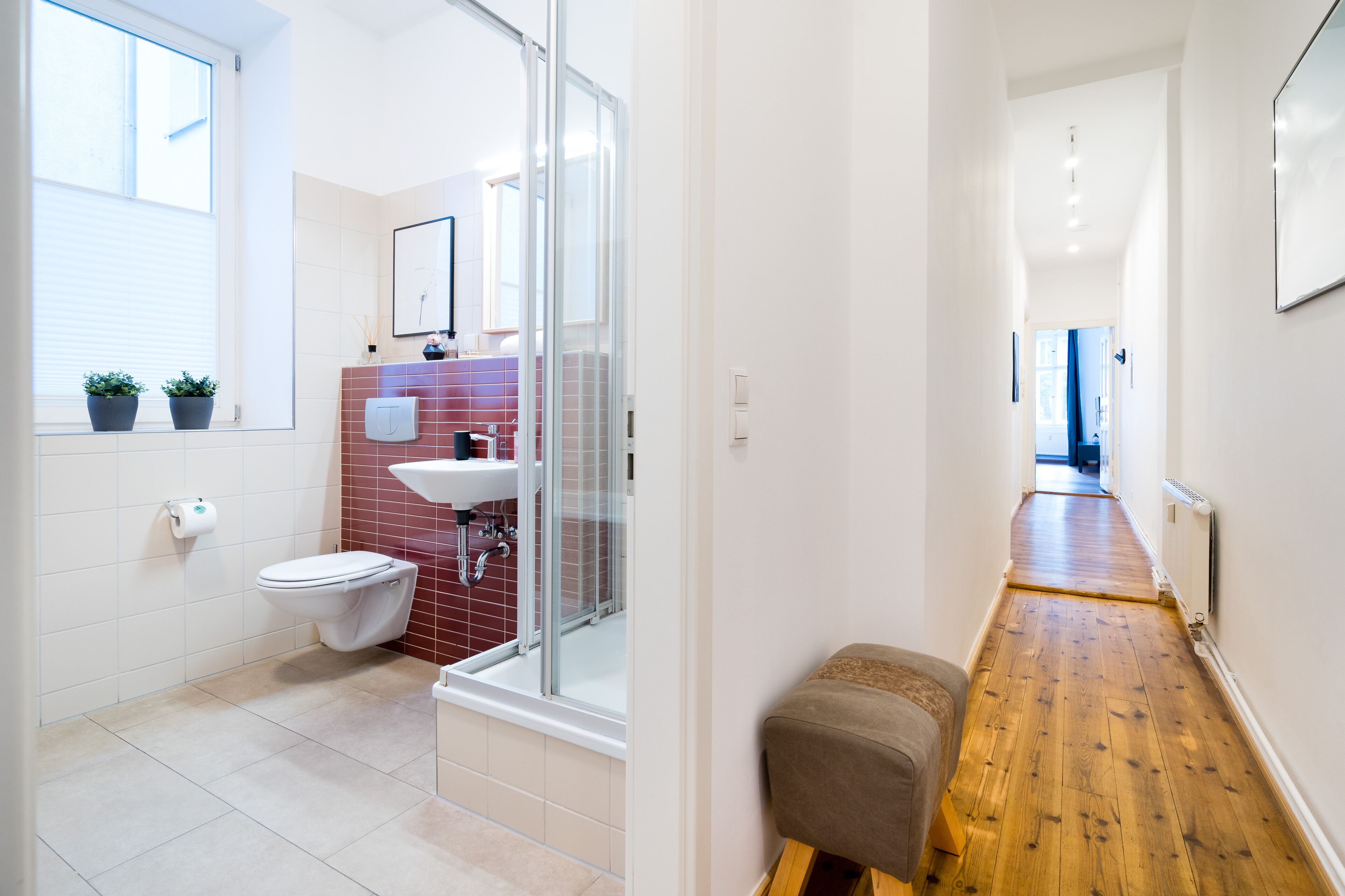 A modern bathroom with a toilet, sink, and shower. There is a wooden bench in the hallway.