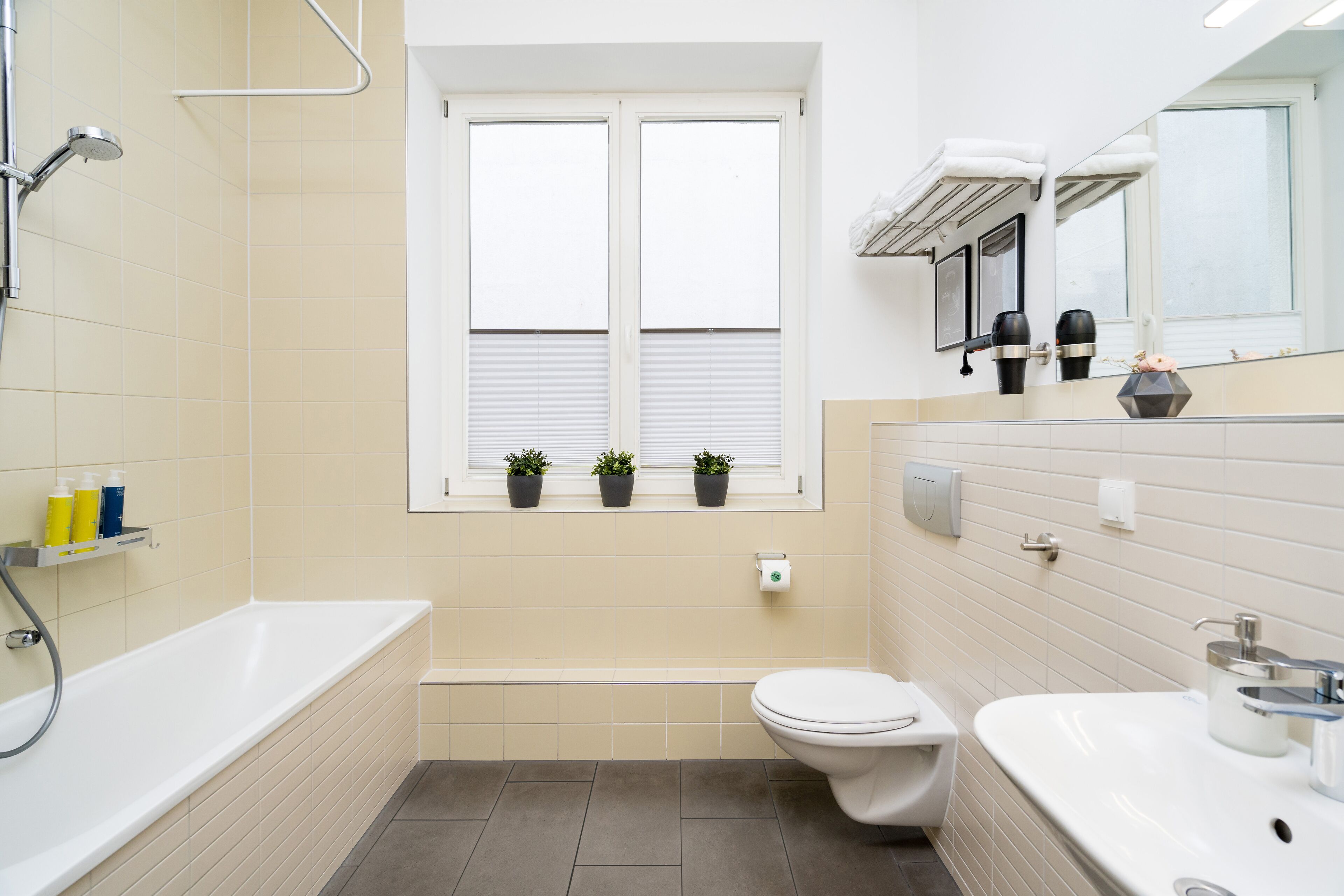 Ein modernes Badezimmer mit Badewanne, Toilette und Waschbecken. Ein Fenster mit Jalousien und ein Ablagefach mit Pflegeprodukten.