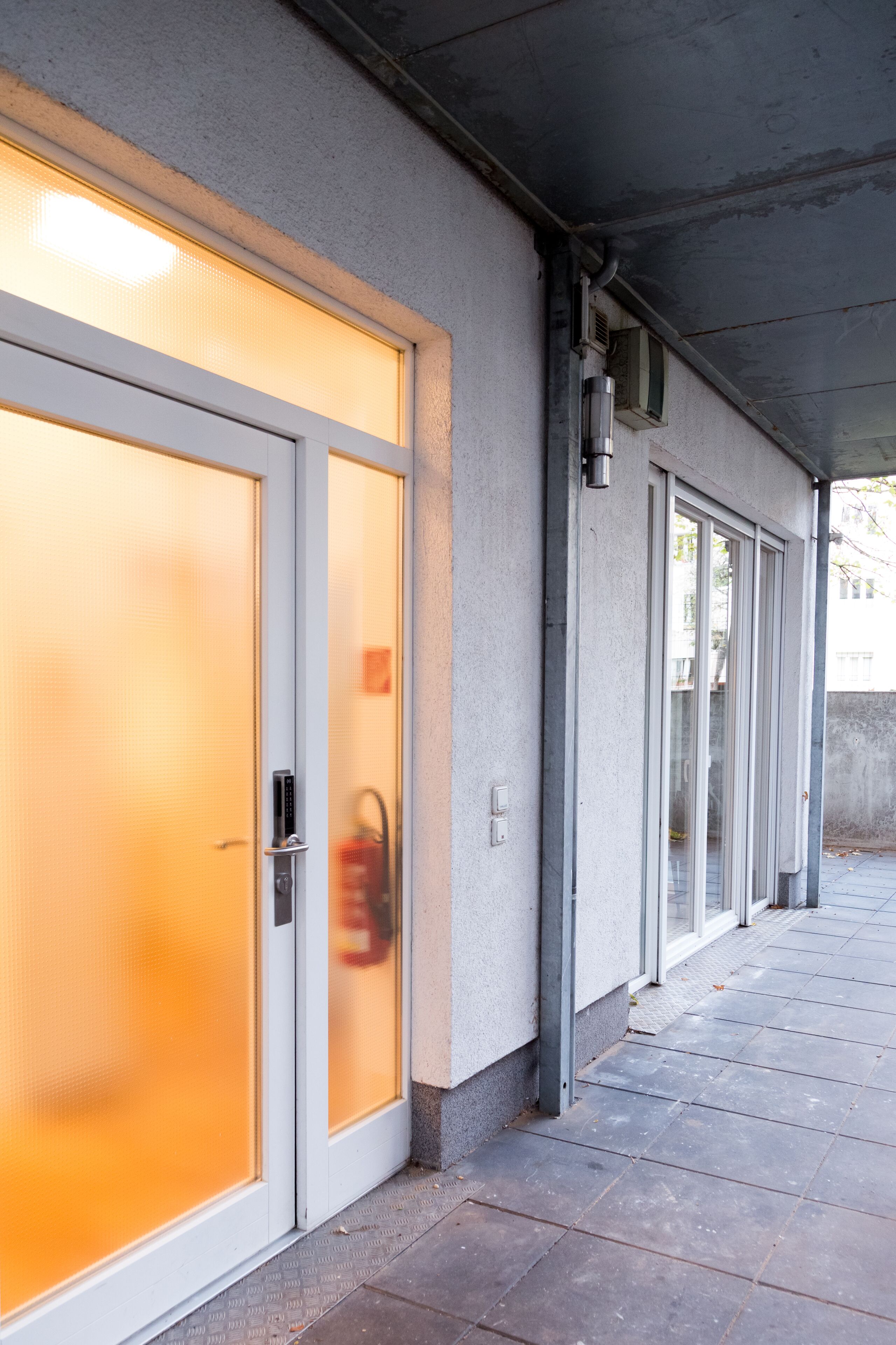 Exterior d'un edifici amb un passadís cobert, portes de vidre i un extintor d'incendis metàl·lic.