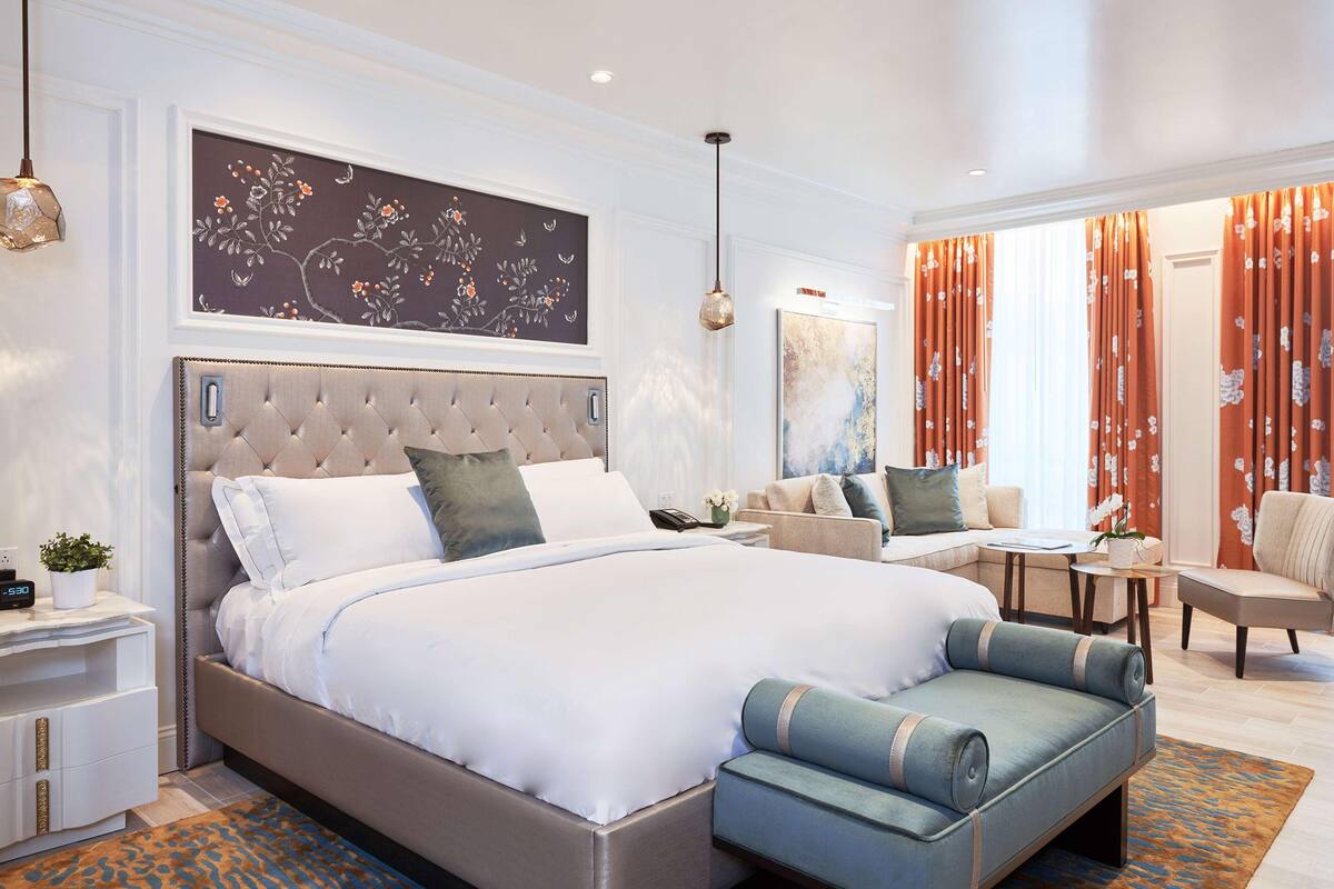 A modern hotel room with a large bed, a sofa, a dining area, and a view of the city.