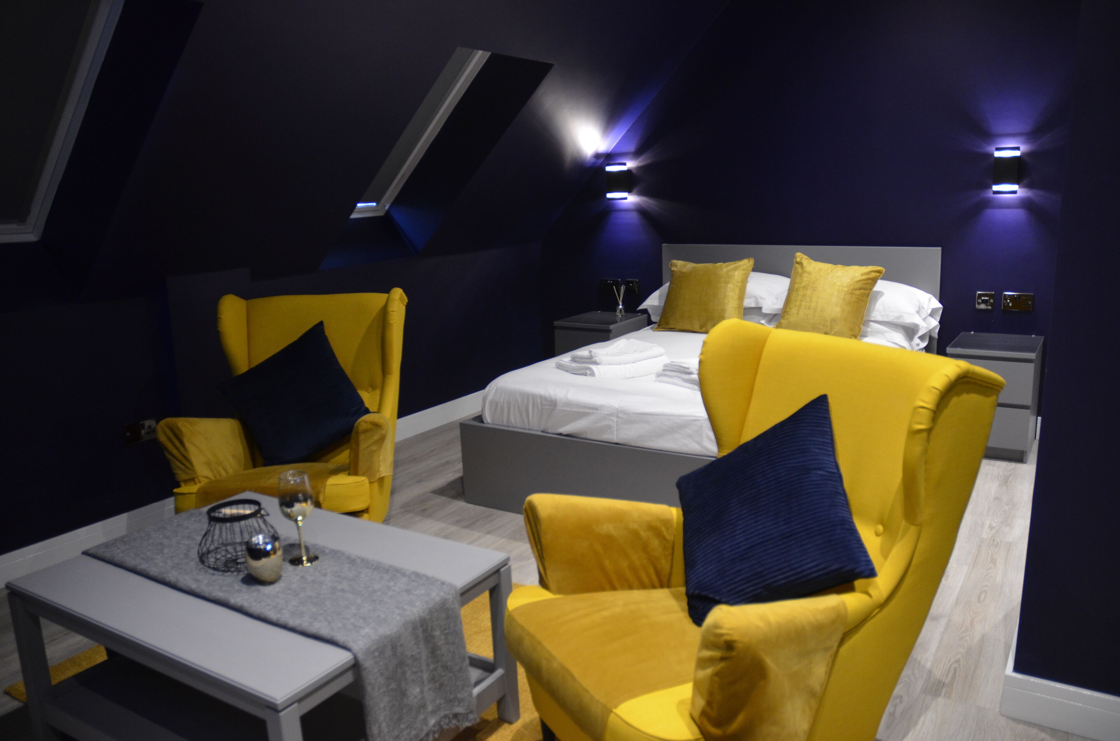 A modern bedroom with a large bed, two yellow armchairs, a small table with a glass and candle, and a skylight above.