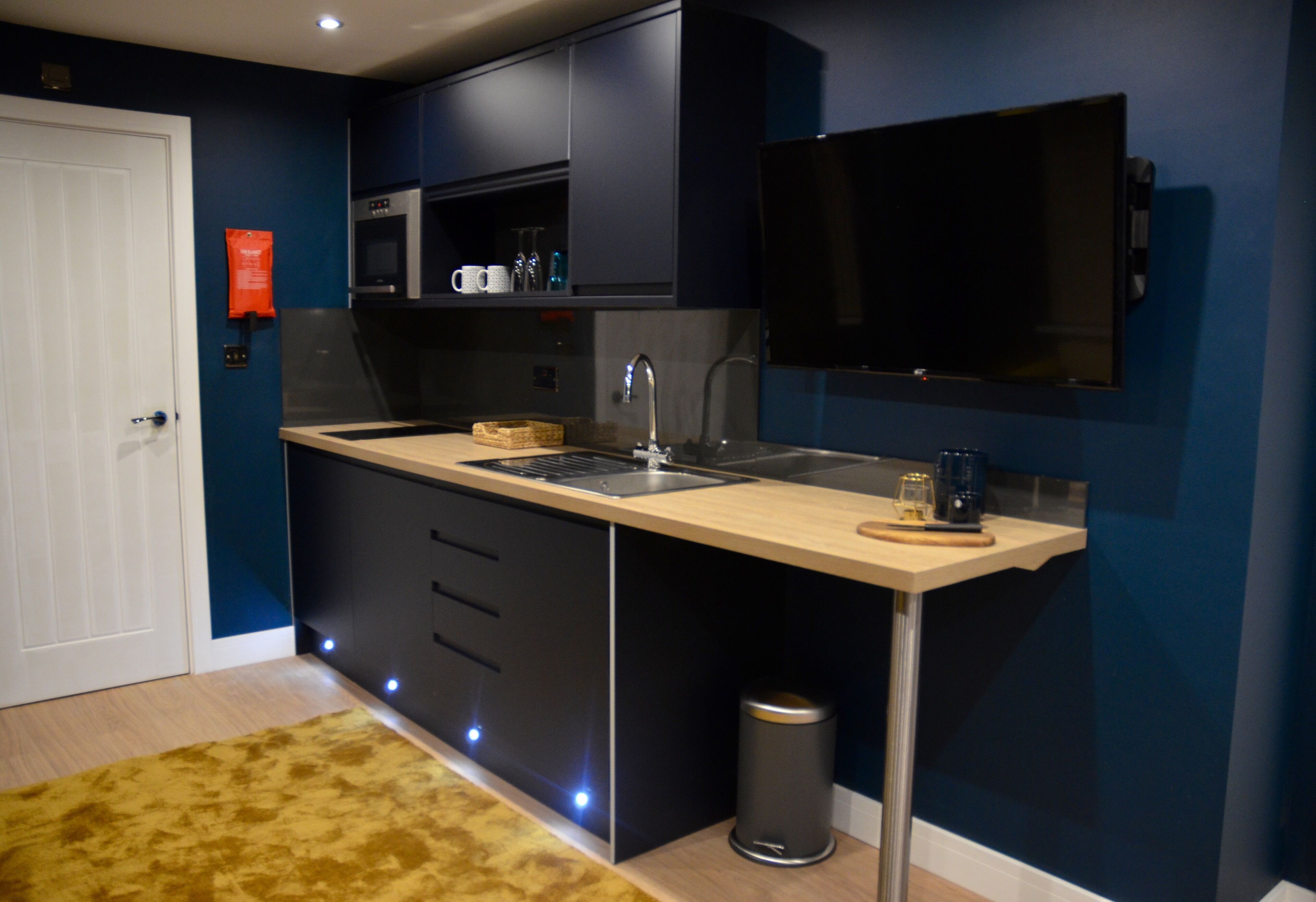 A modern kitchen with a built-in oven, microwave, and sink. There is a trash can and a small rug on the floor.