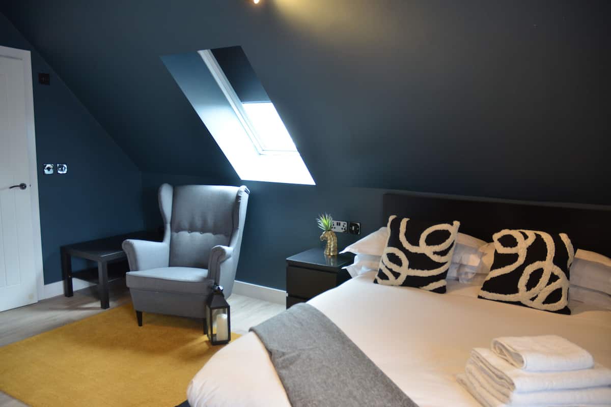 Une chambre avec un puits de lumière, un lit avec des oreillers blancs et à motifs, un fauteuil gris, une petite table avec une lanterne et un tapis jaune.