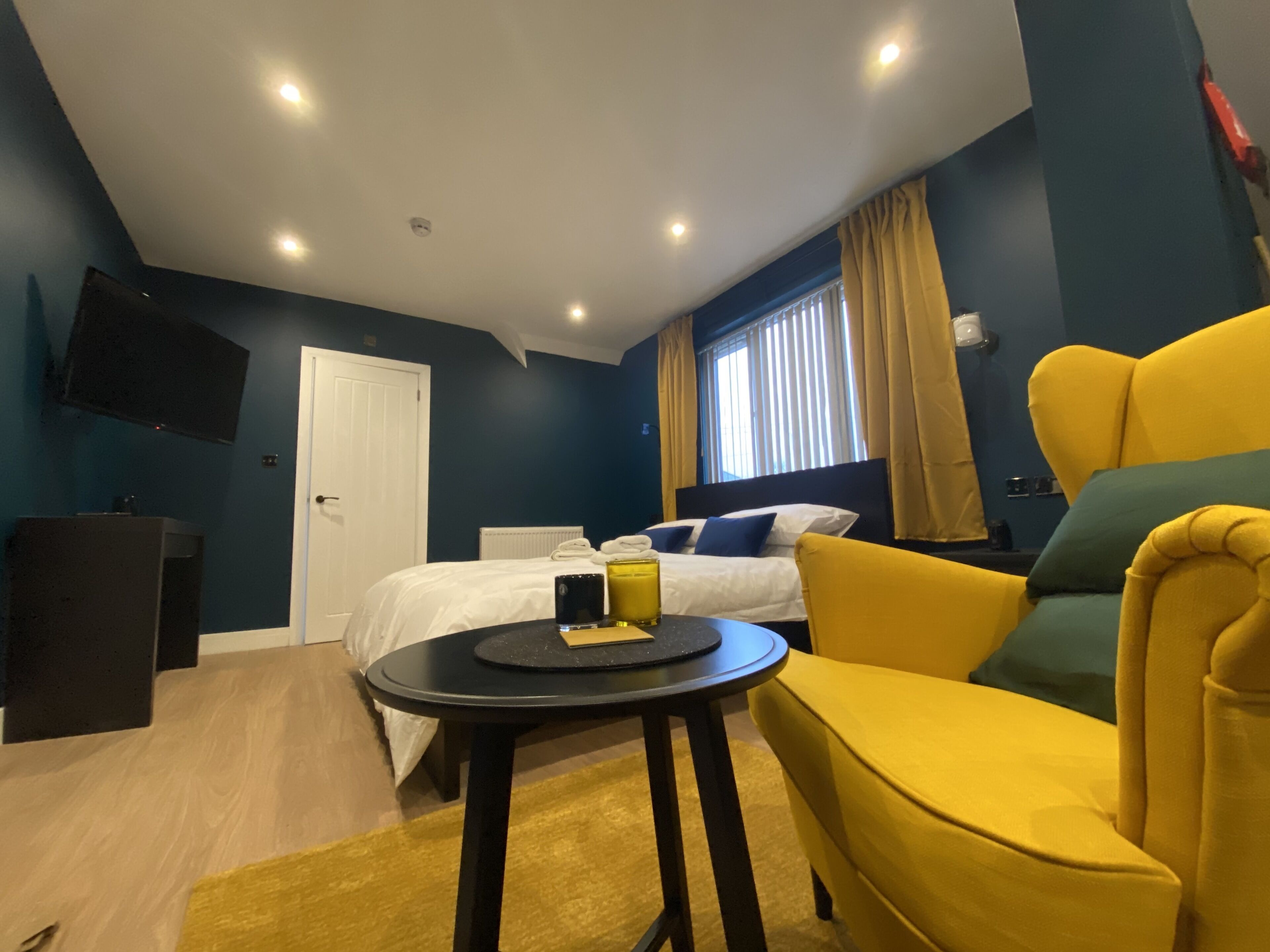 A modern hotel room with a bed, a yellow armchair, a small round table, and a TV mounted on the wall.