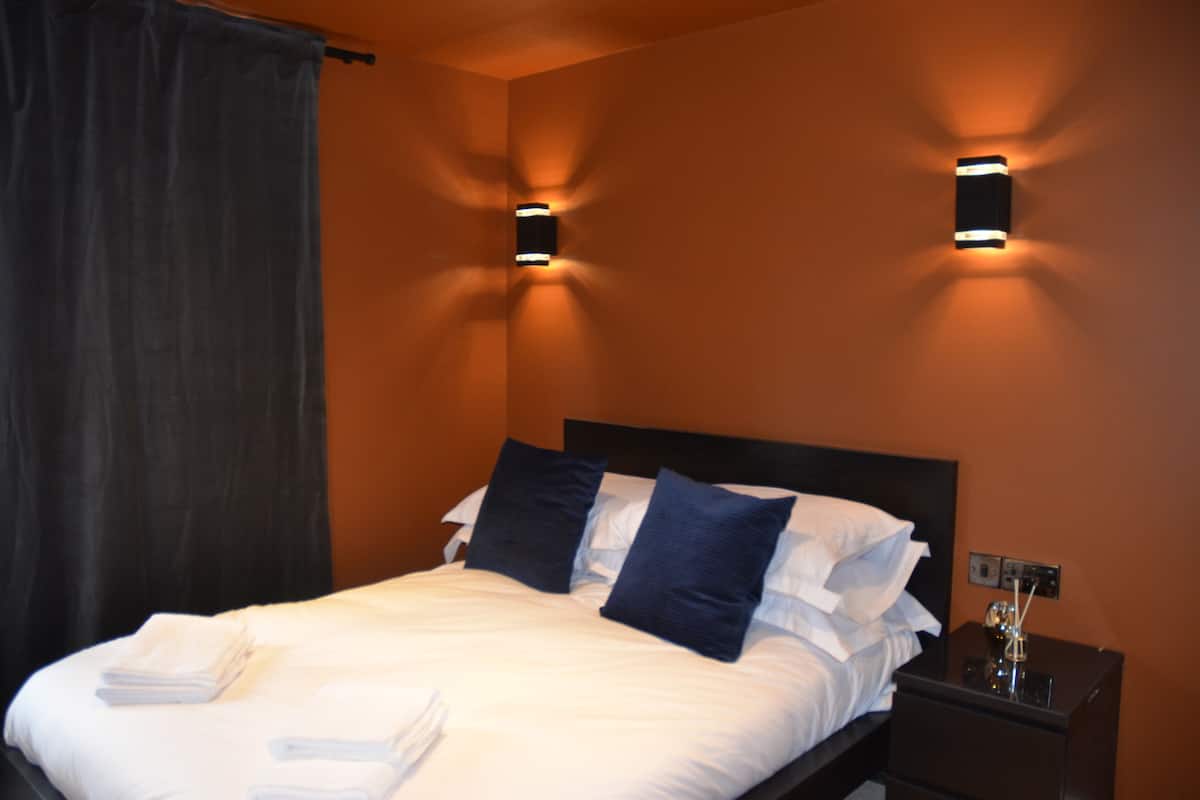 Un lit bien fait, avec du linge de lit blanc et deux oreillers bleu foncé, une table de chevet noire avec une lampe, et deux appliques murales fixées sur un mur orange.
